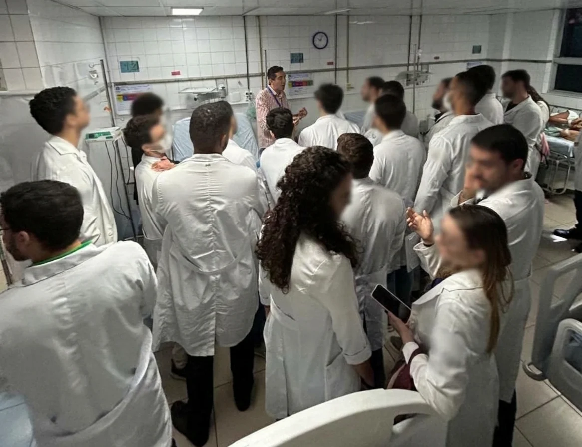 Estudantes de jaleco reunidos em volta do leito em uma enfermaria, acompanhando a explicação do professor